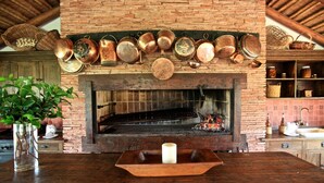 A rustic kitchen with a brick fireplace, hanging copper pots, and a wooden table with a candle.