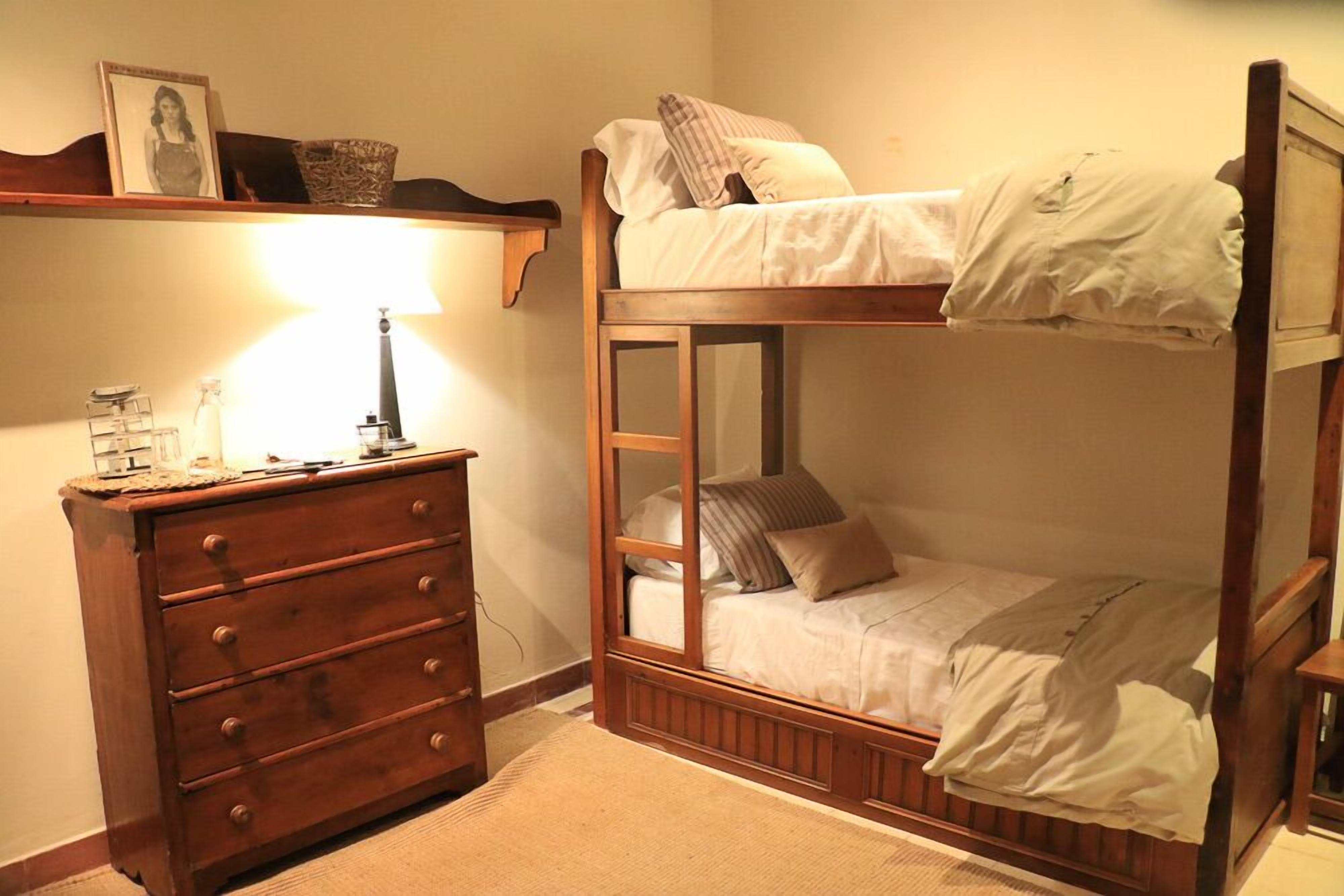 Una cama litera con estructura de madera, un aparador con cajones, una lámpara y un cuadro en la pared.