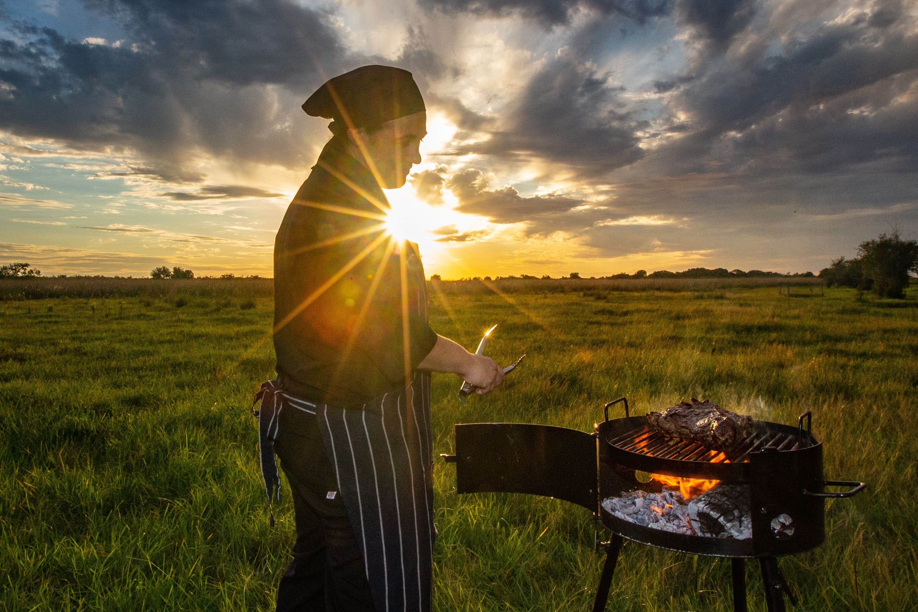 A person grilling outdoors with a sunset in the background.