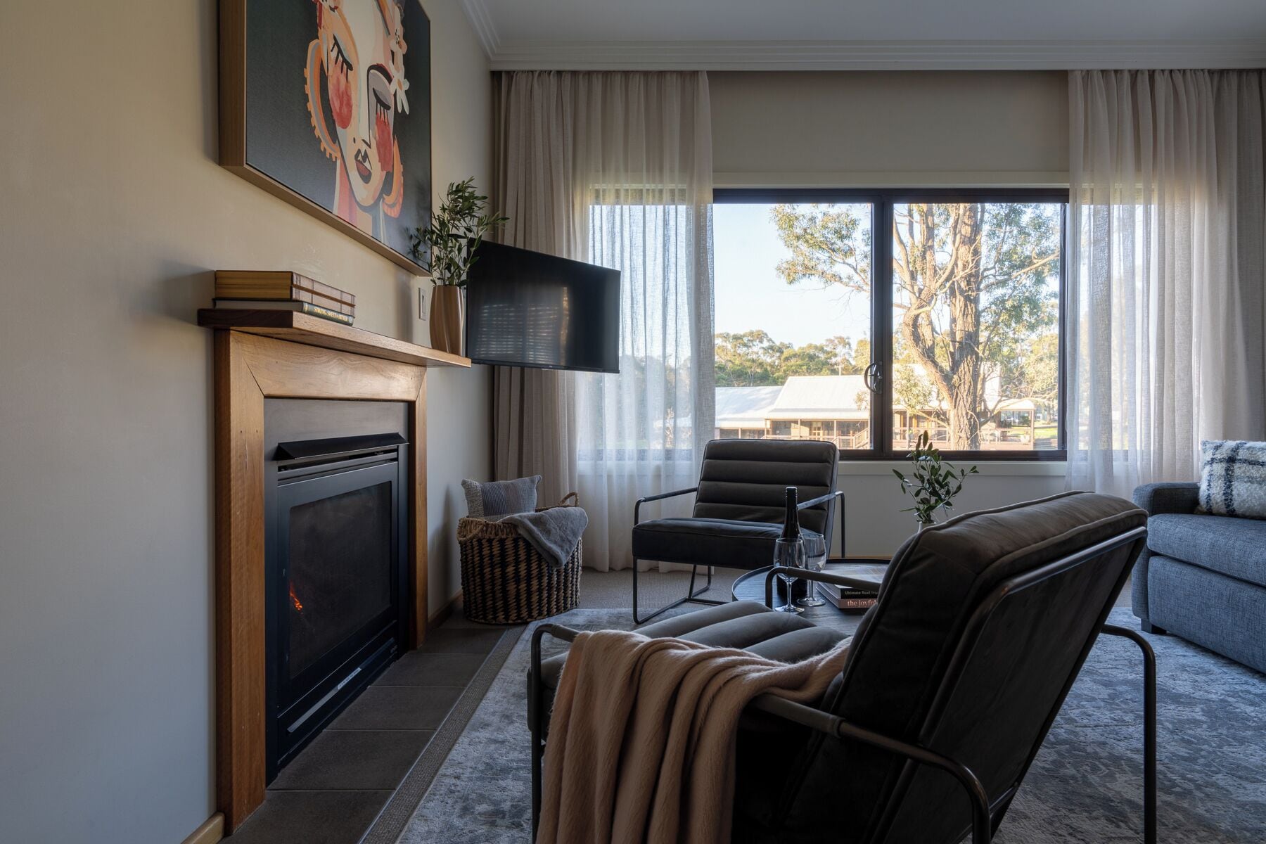 Una sala de estar con chimenea, televisor de pantalla plana, zona de estar acogedora y un ventanal con vistas a árboles y edificios.