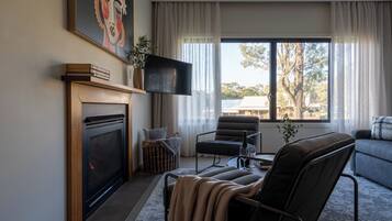 Una sala de estar con chimenea, televisor de pantalla plana, zona de estar acogedora y un ventanal con vistas a árboles y edificios.
