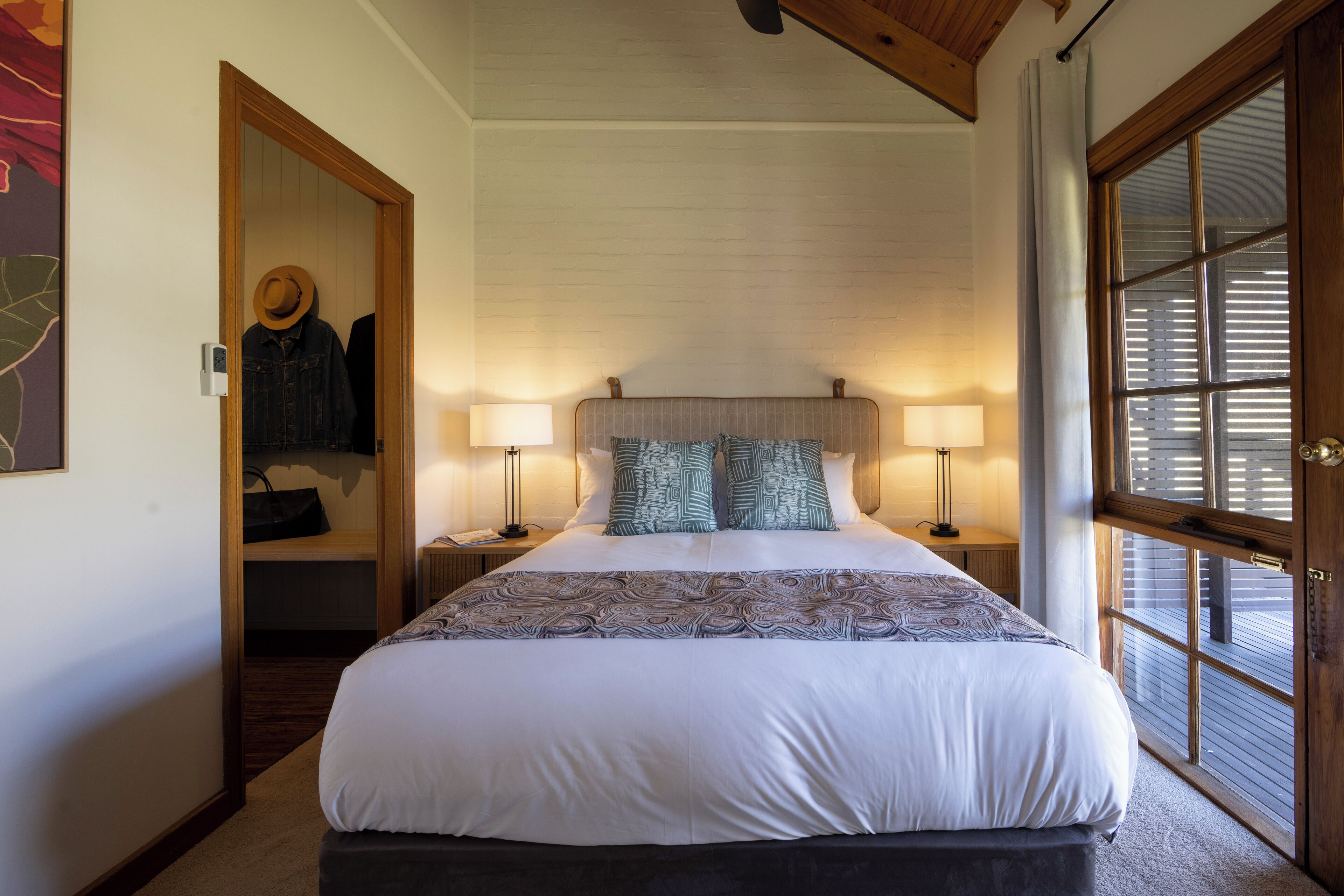 A neatly made bed with patterned bedding, two bedside lamps, and a view through a window with blinds.