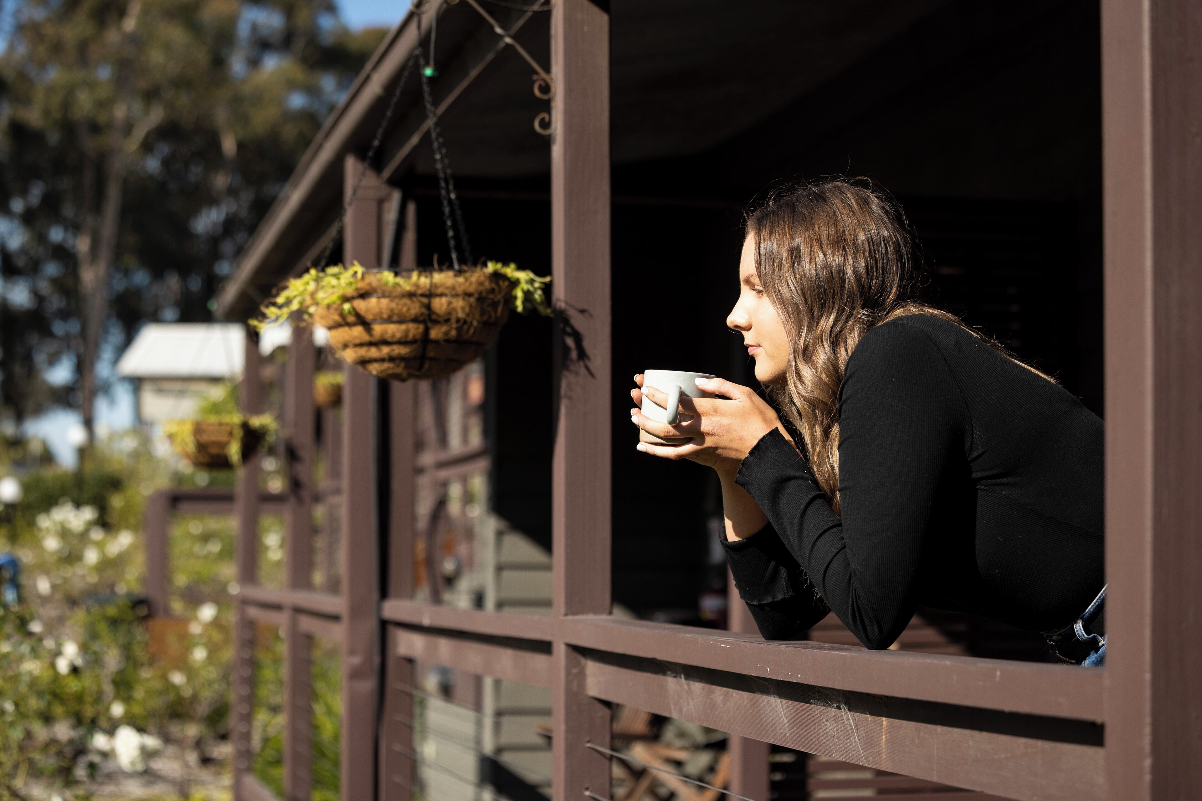 Una persona sentada junto a una ventana con una planta colgante, sosteniendo una taza.