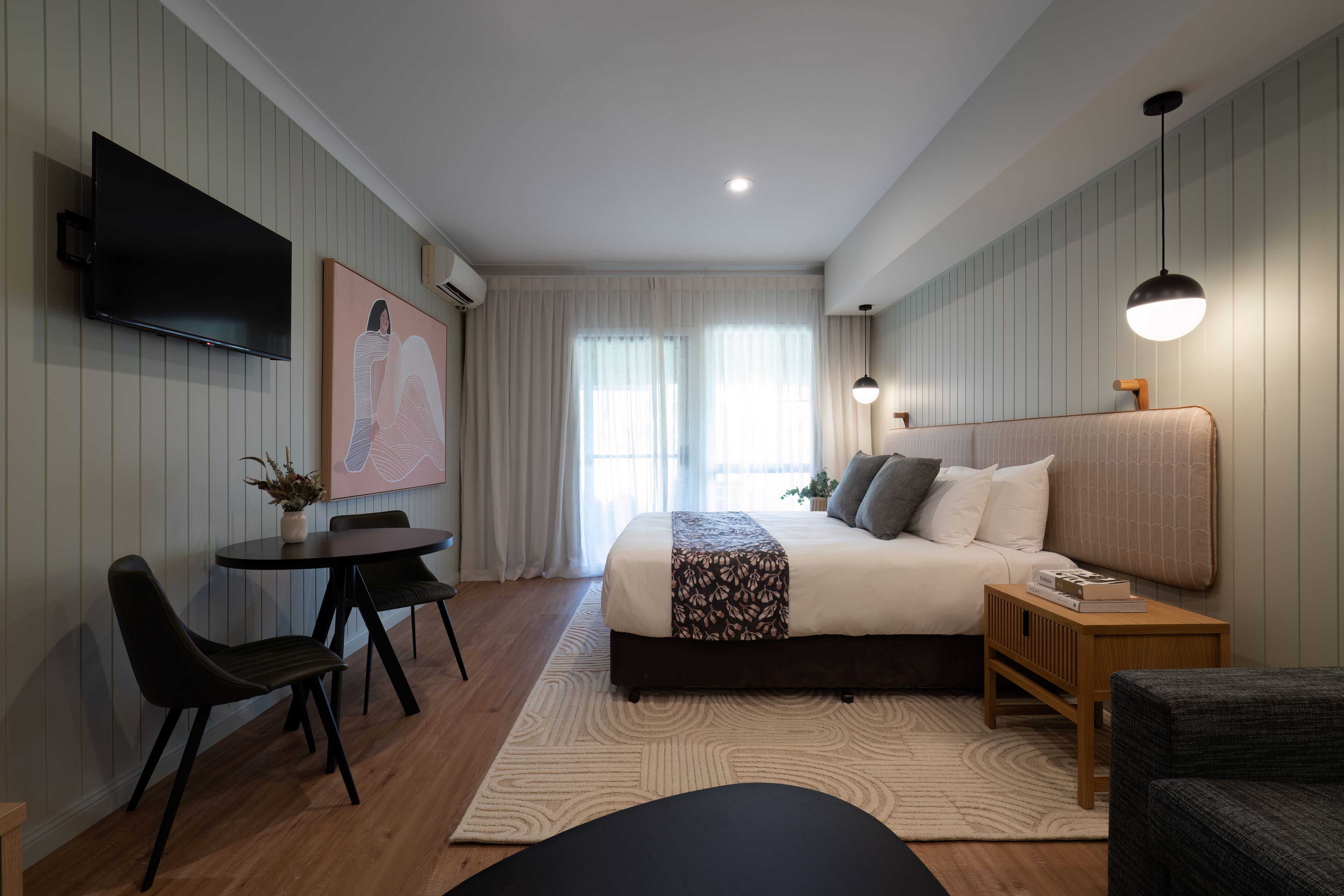 A modern bedroom with a large bed, a TV mounted on the wall, a small dining table with chairs, and a bedside table with a lamp.