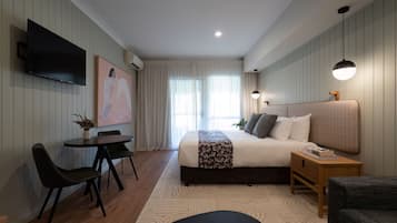 Un dormitorio moderno con una cama grande, un televisor montado en la pared, una pequeña mesa de comedor con sillas y una mesita de noche con una lámpara.