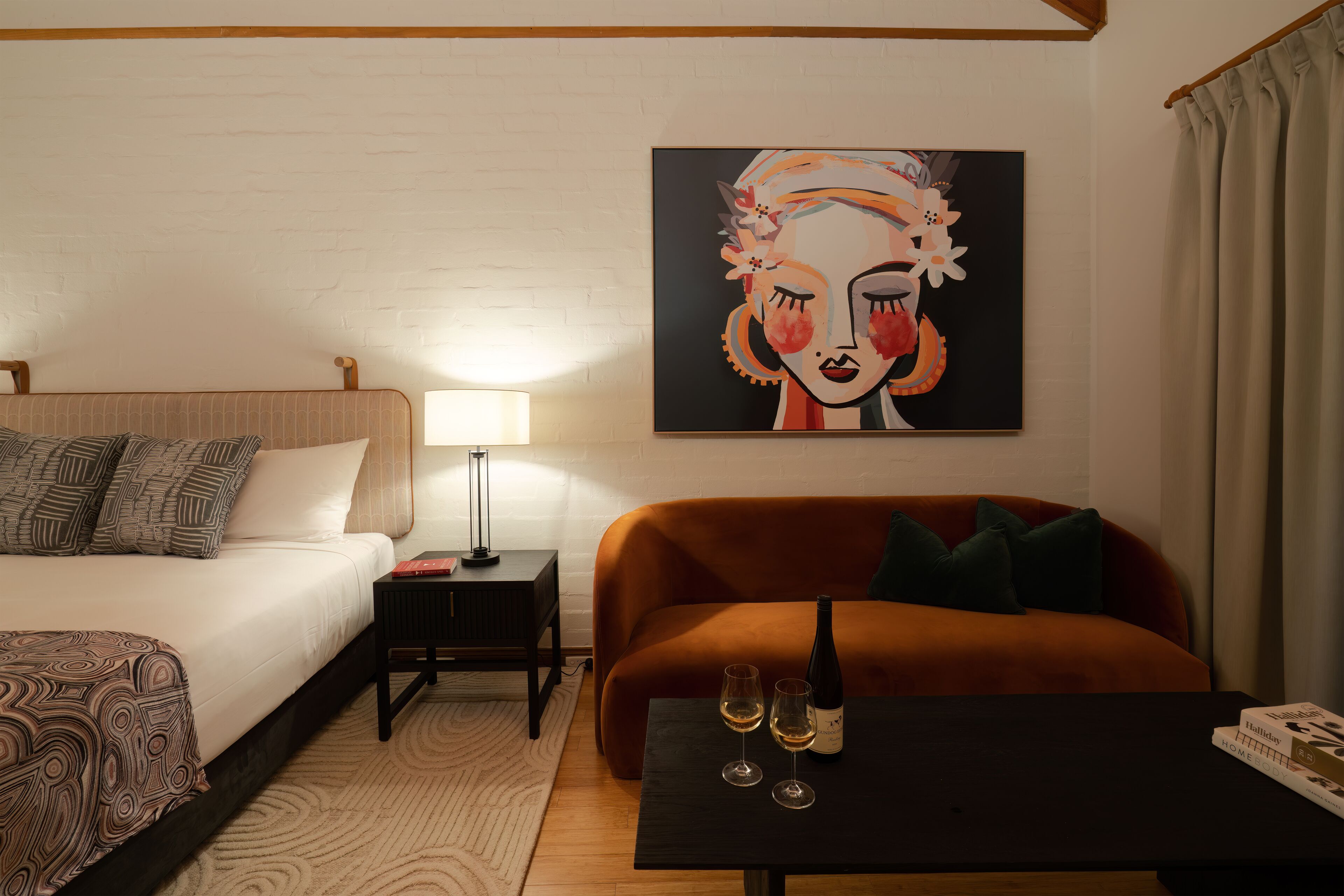 Een moderne hotelkamer met een bed, een bank, een klein tafeltje met een fles en glazen, en een schilderij aan de muur.