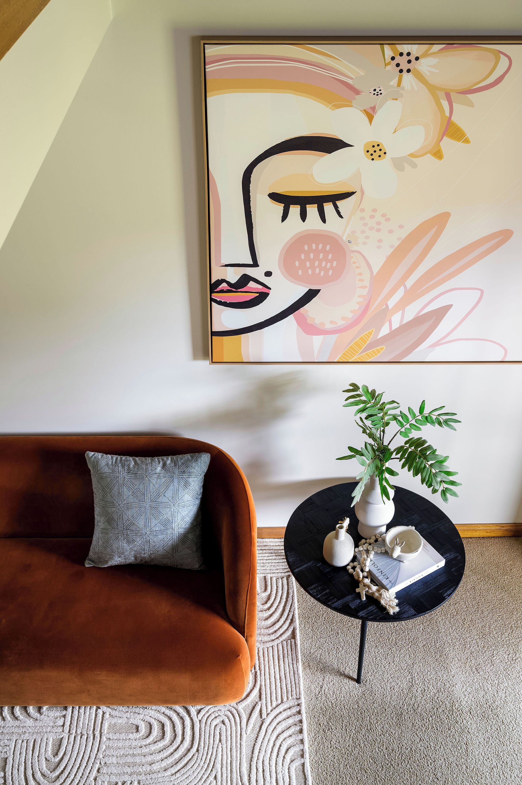 A living room with a brown sofa, a patterned throw pillow, a round black side table with a white vase and decorative items, and a large abstract painting on the wall.