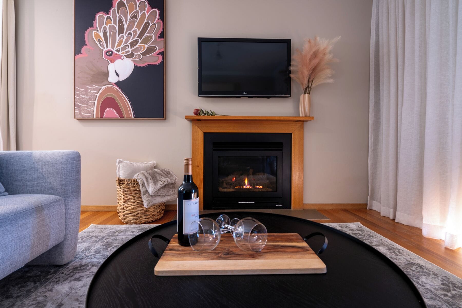 A living room with a fireplace, a flat-screen TV, a sofa, a decorative wall piece, and a bottle of wine on a tray.