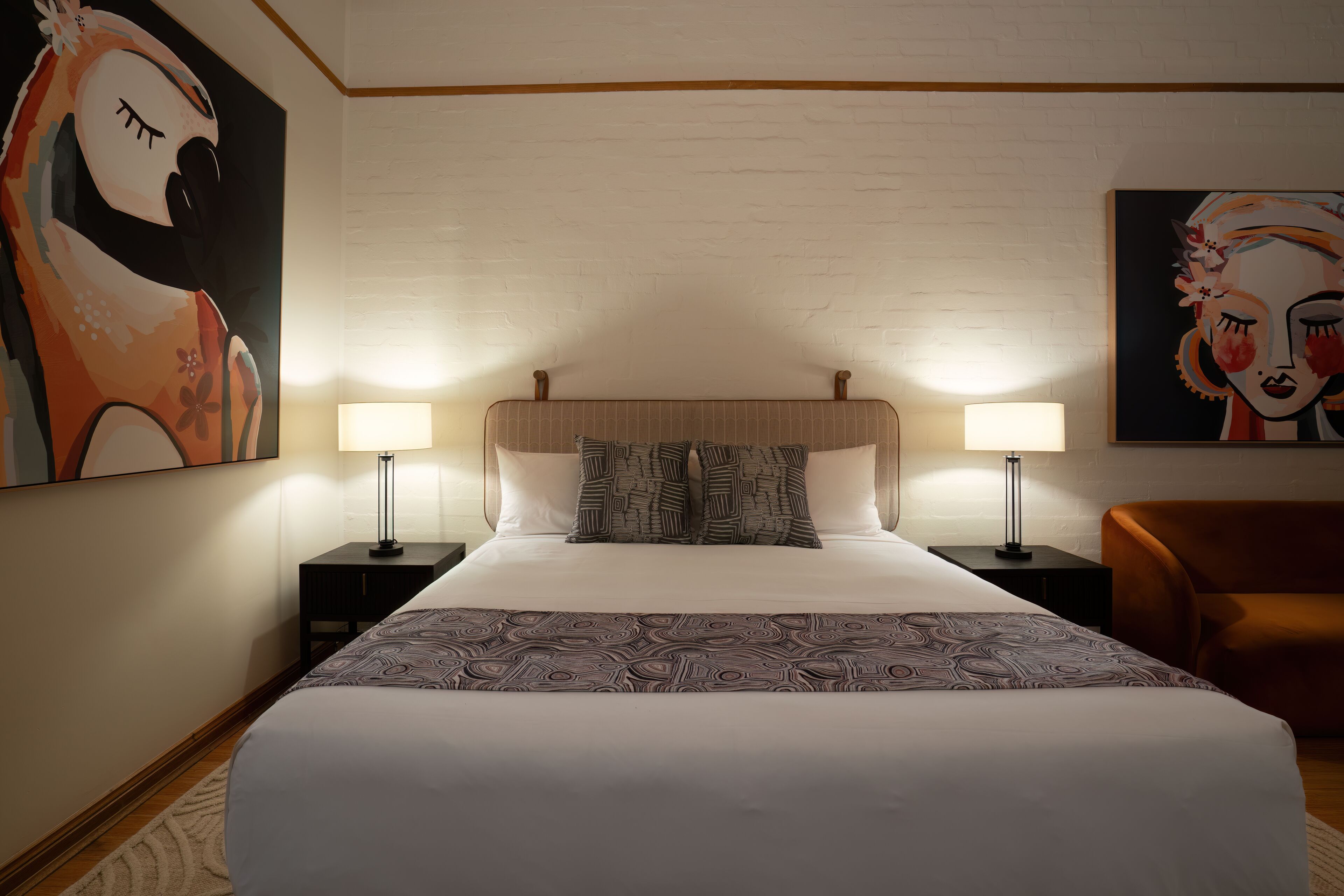 A hotel room with a large bed, two bedside tables with lamps, a brown armchair, and two paintings on the walls.