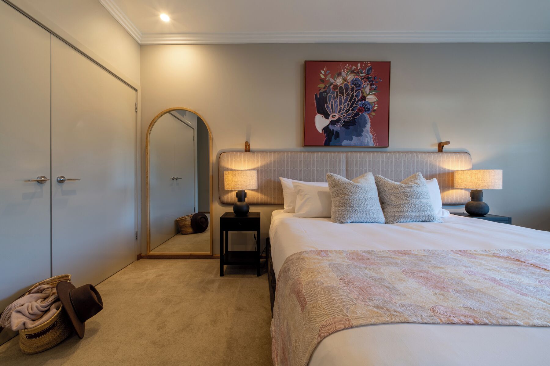 A hotel room with a large bed, two bedside lamps, a mirror, a painting above the bed, and a wardrobe.