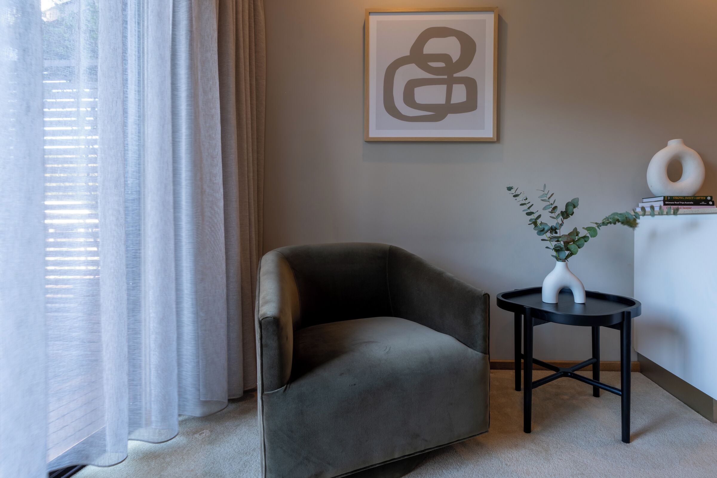 A modern living room with a grey armchair, a small round table with a vase and plant, and a framed abstract artwork on the wall.