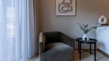 A modern living room with a grey armchair, a small round table with a vase and plant, and a framed abstract artwork on the wall.