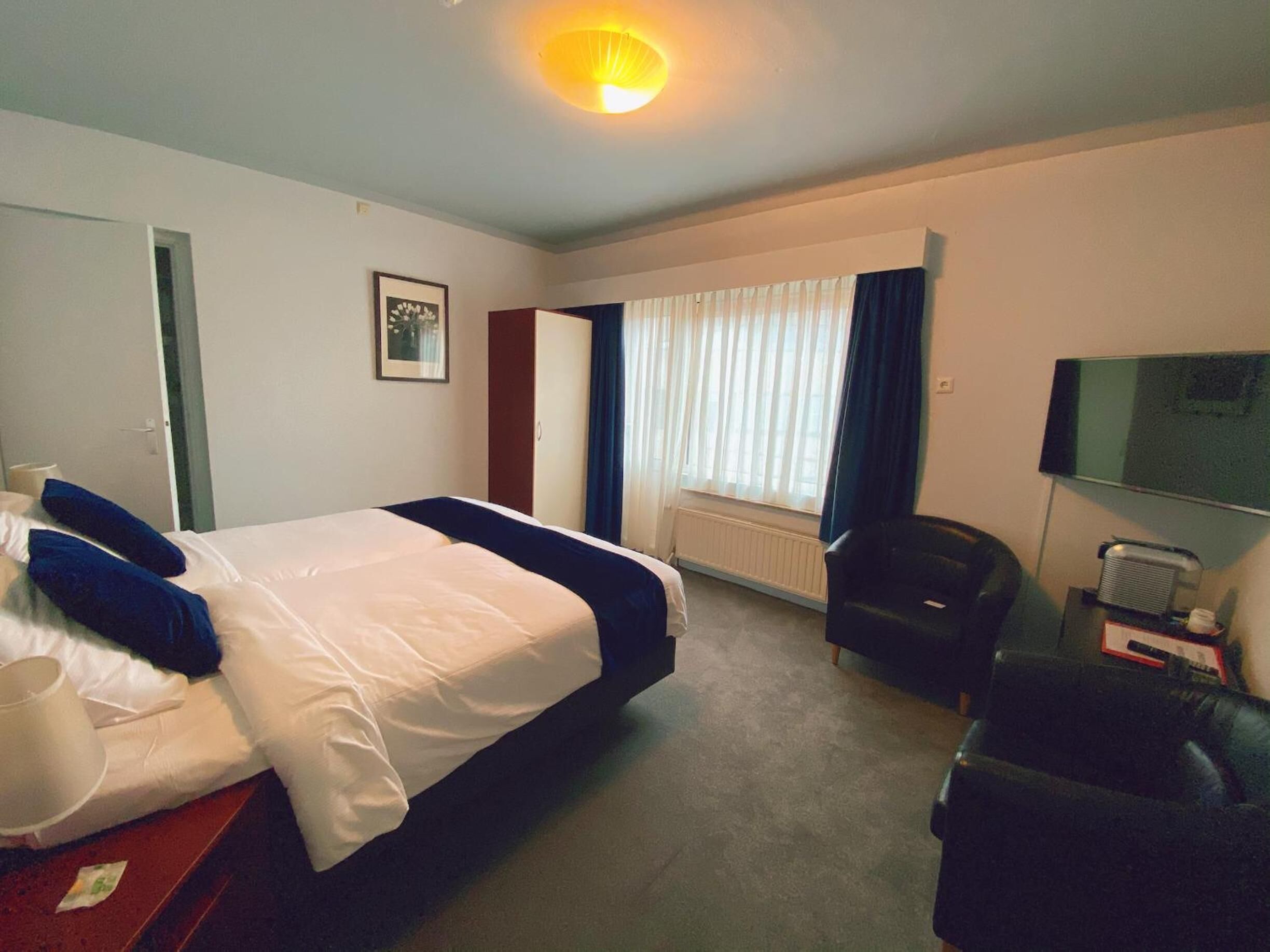 Una habitación de hotel con cama, una silla, un escritorio con computadora, televisor y ventana con cortinas.