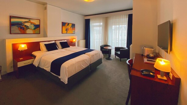 Ein Hotelzimmer mit einem großen Bett, zwei Nachttischen inklusive Lampen, einem Schreibtisch mit Stuhl, einem Flachbildfernseher und Bildern an den Wänden.