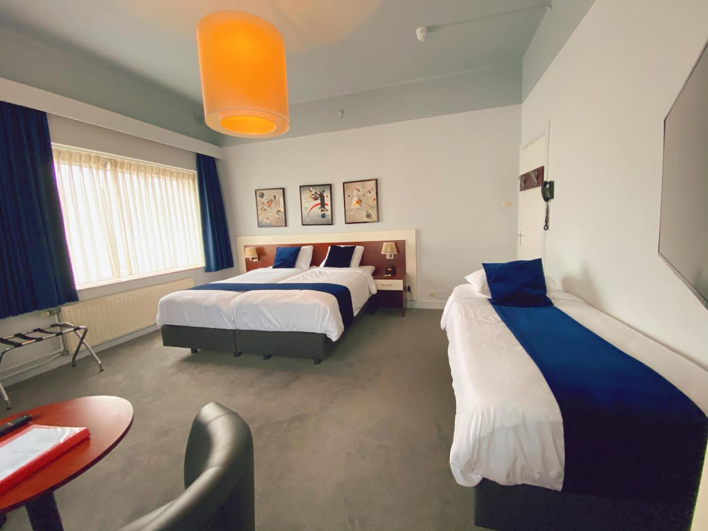 A hotel room with two beds, a desk, and a chair. There are three paintings on the wall, a window with curtains, and a ceiling light.