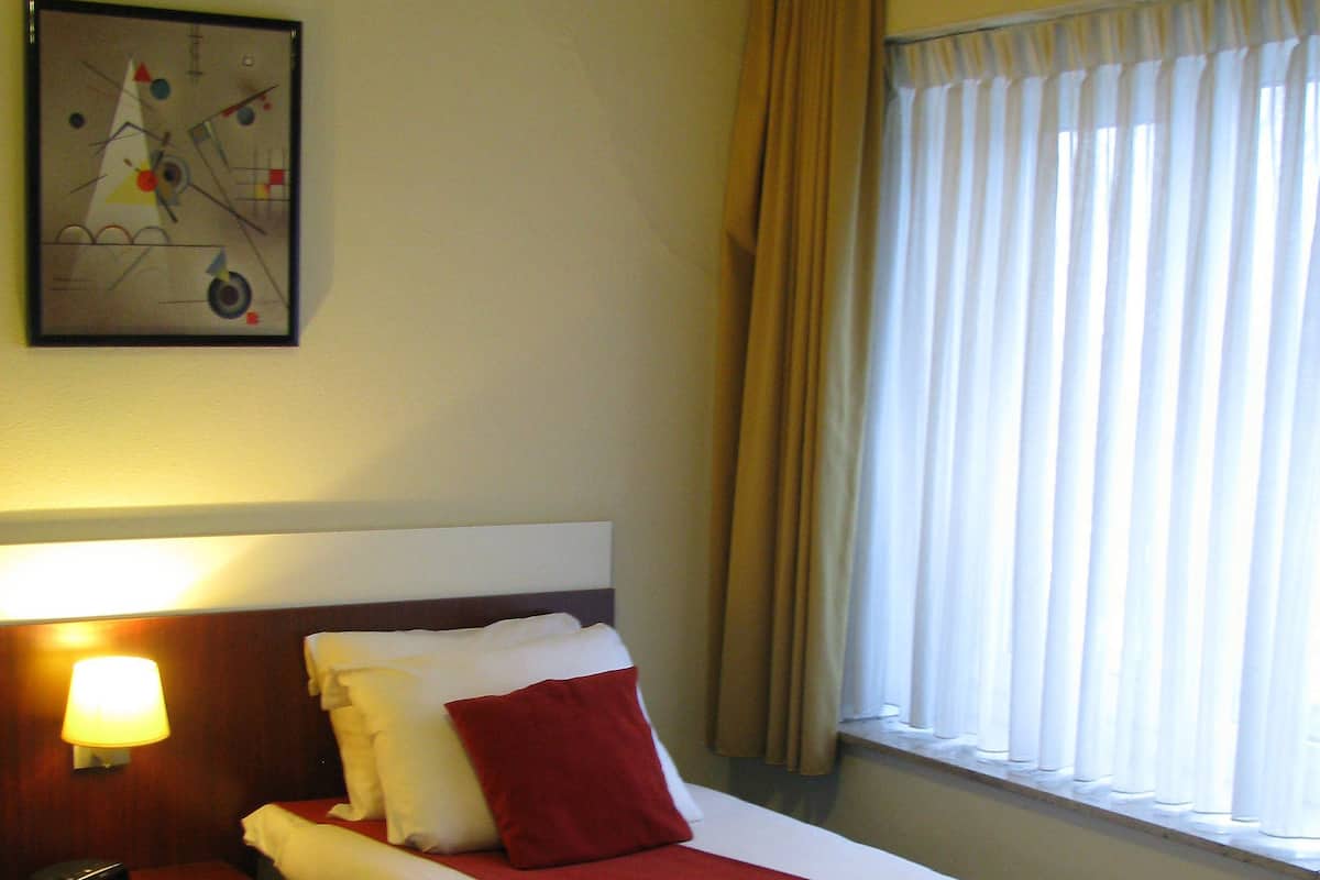 A hotel room with a single bed, a nightstand with a lamp, a wall clock, and a window with curtains.
