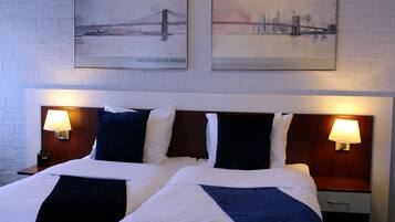 Habitación de hotel con dos camas, cada una con cabecero, y dos lámparas fijadas a la pared. Dos cuadros enmarcados decoran la pared sobre las camas.