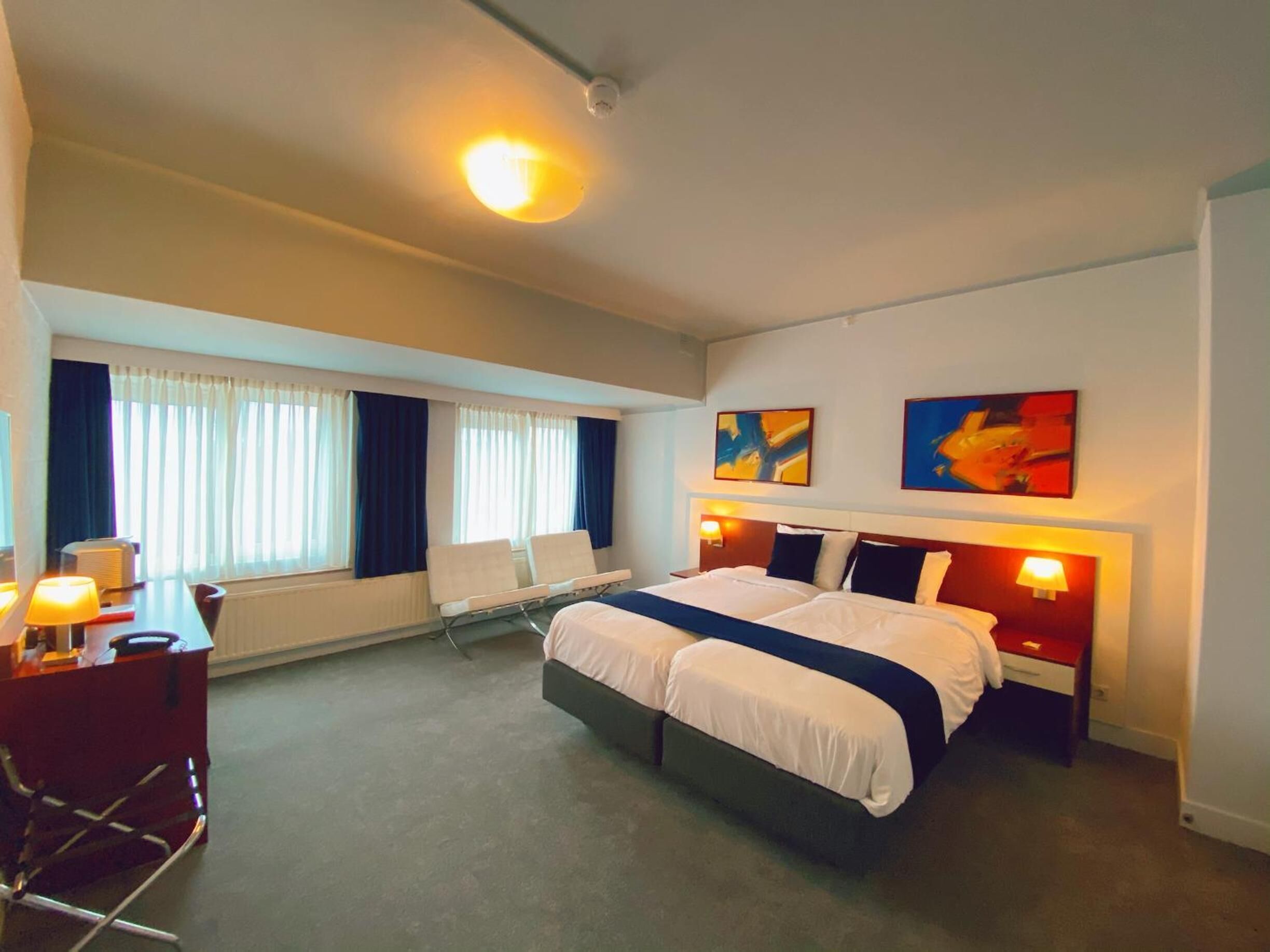 A hotel room with a large bed, a desk with a chair, a window with curtains, and two abstract paintings on the wall.