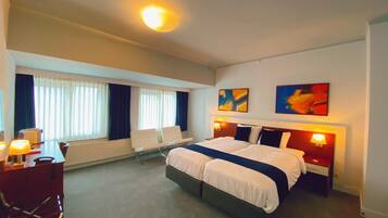 A hotel room with a large bed, a desk with a chair, a window with curtains, and two abstract paintings on the wall.