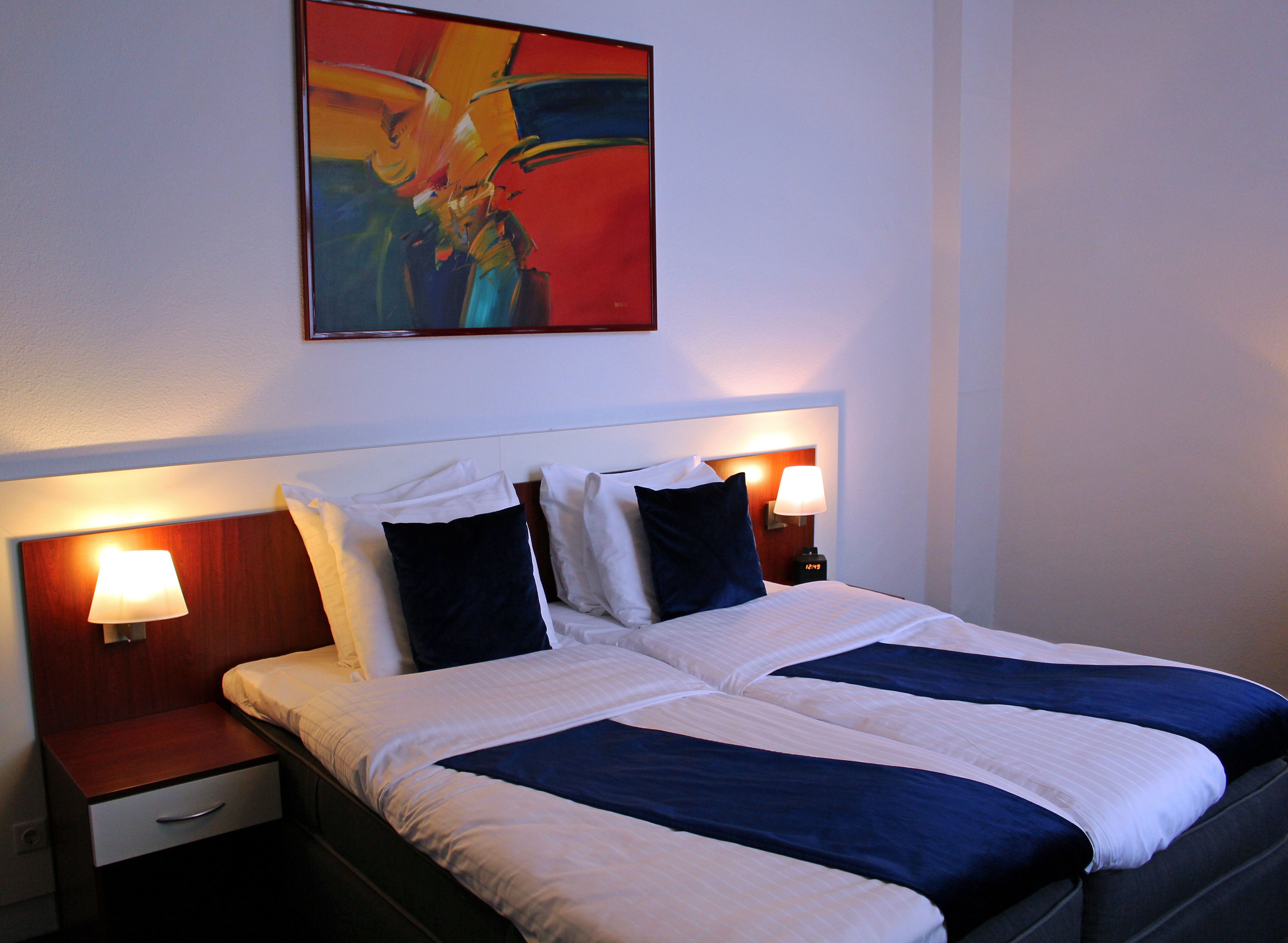 A double bed with white and navy bedding, two bedside tables with lamps, and a painting above the bed.