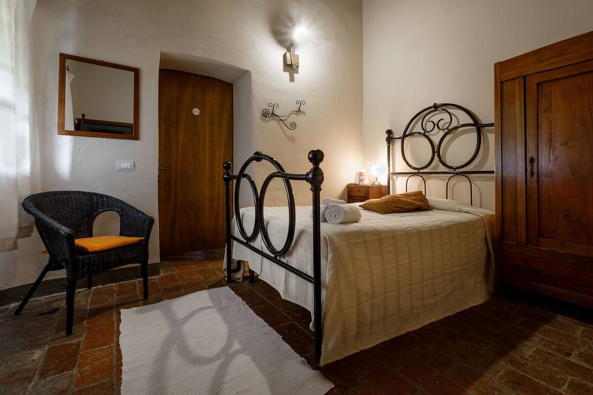 A bedroom with a four-poster bed, a wicker chair, a wooden wardrobe, and a mirror on the wall.