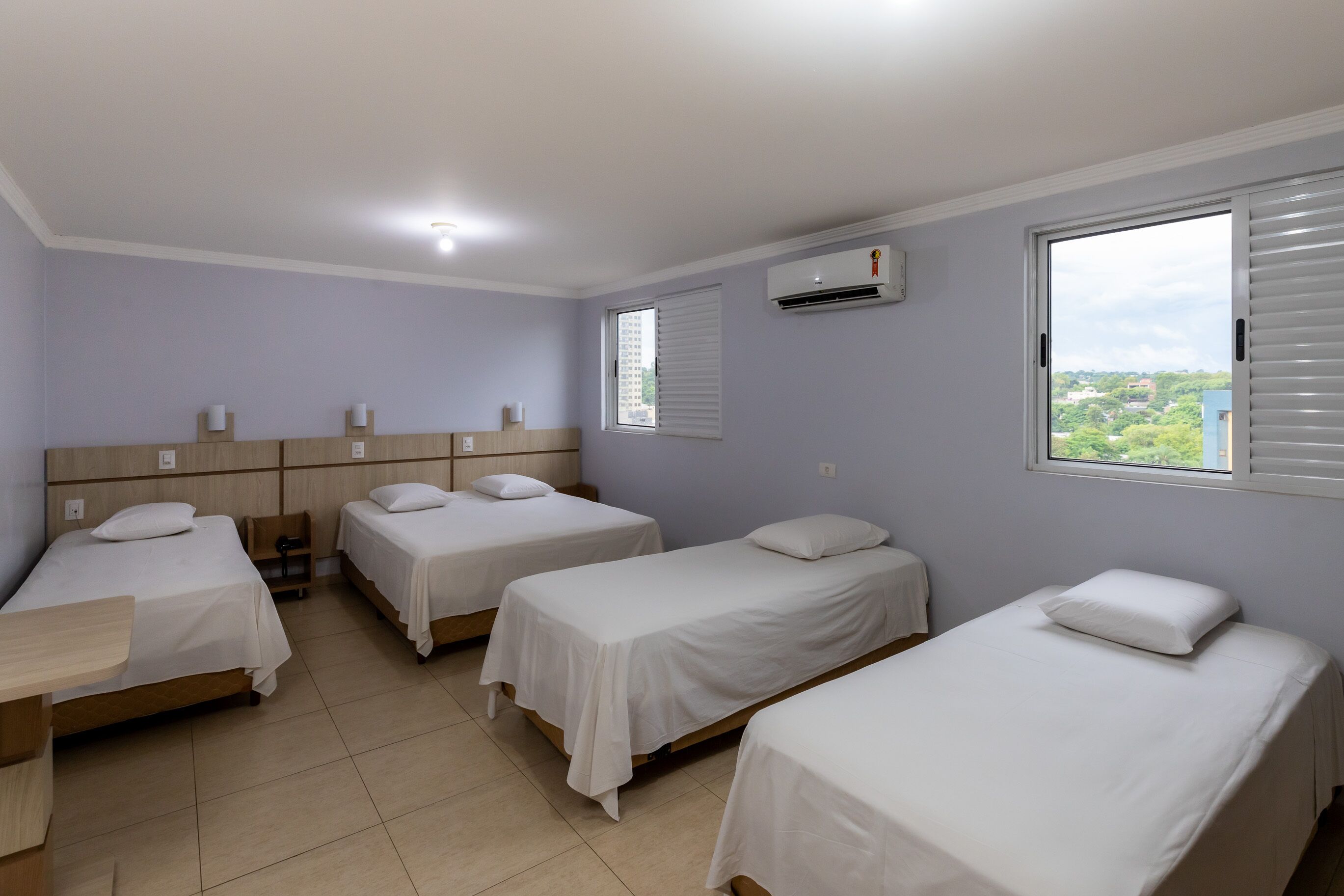 Ein Hotelzimmer mit vier Einzelbetten, einem Holztisch, einem Fenster mit Jalousien und einer Klimaanlage.