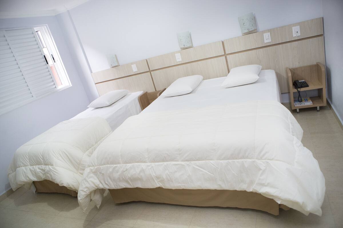 Een hotelkamer met twee bedden, een nachtkastje, een raam met zonwering en een houten hoofdeinde.