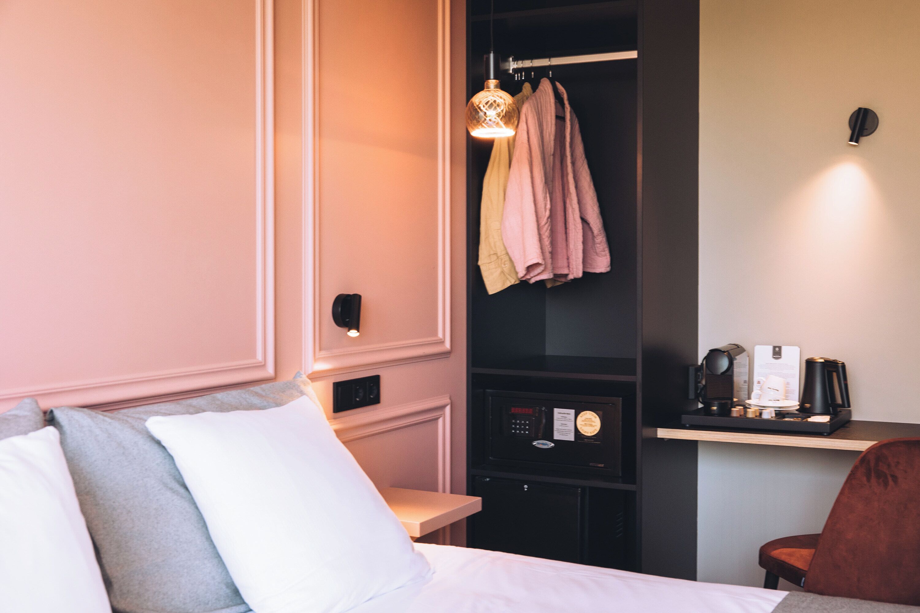 Camera d'albergo con un letto, cuscini, un armadio, uno scaffale con una macchina per il caffè e una lampada a parete.