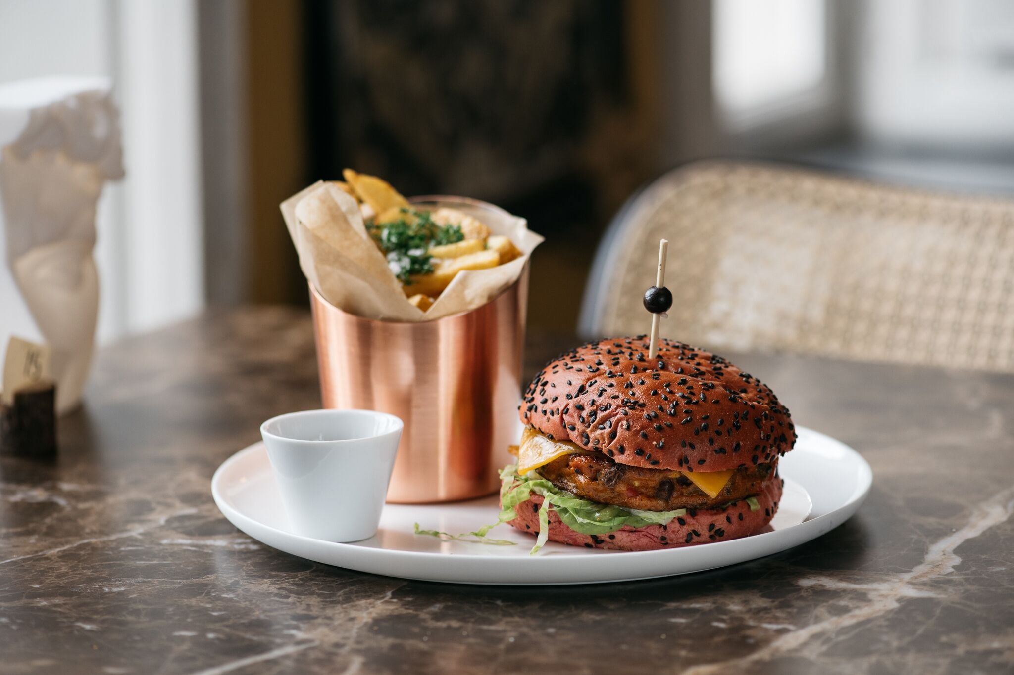 A burger with a sesame seed bun, a side of fries, and a small cup on a marble table.
