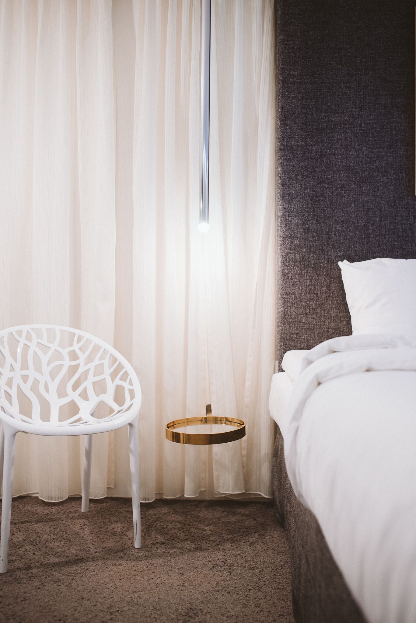 Una habitación de hotel con una cama, una silla blanca, una mesita redonda y cortinas traslúcidas.