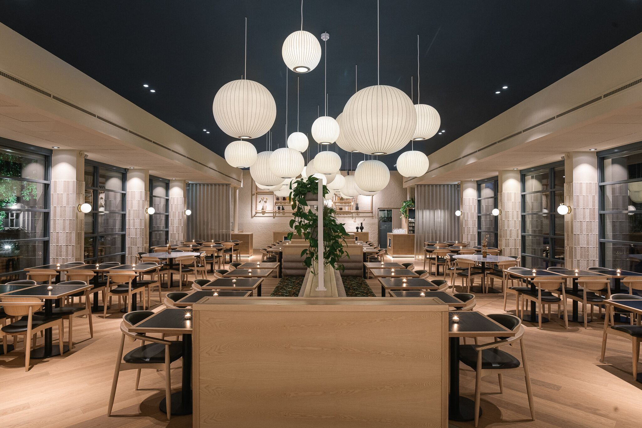 A spacious dining area with multiple tables and chairs, a central plant, and hanging spherical light fixtures.