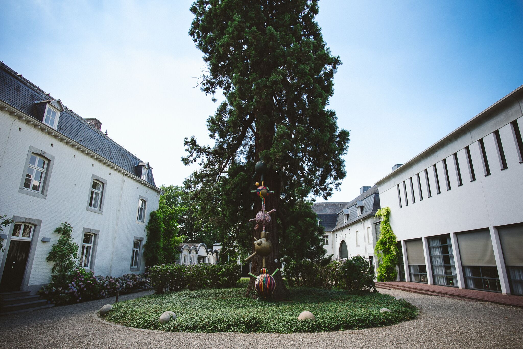 Une cour avec un grand arbre, une sculpture décorative et des bâtiments dotés de fenêtres et de portes.
