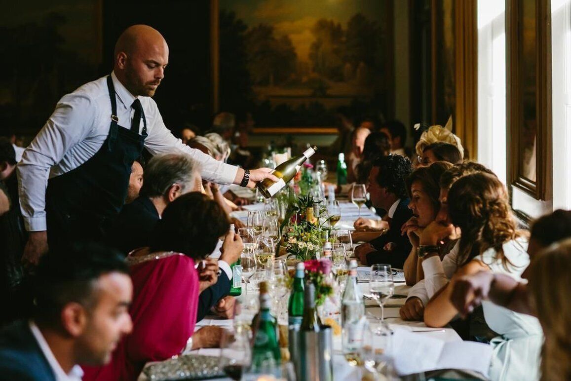A formal dining setting with a long table, guests seated, and a waiter serving wine.
