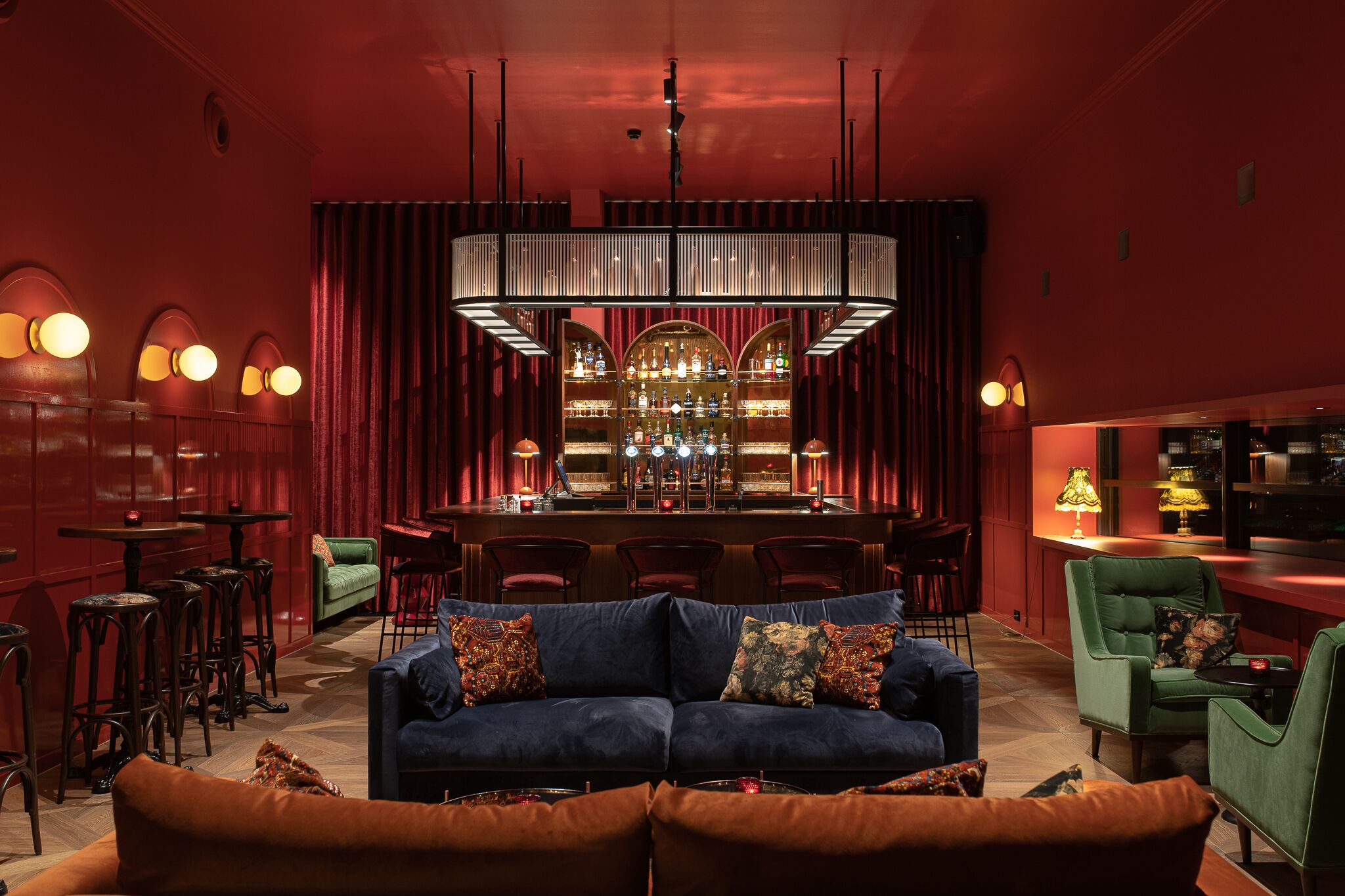 Zona de bar con un fondo rojo, un sofá azul con cojines estampados y una barra con botellas.