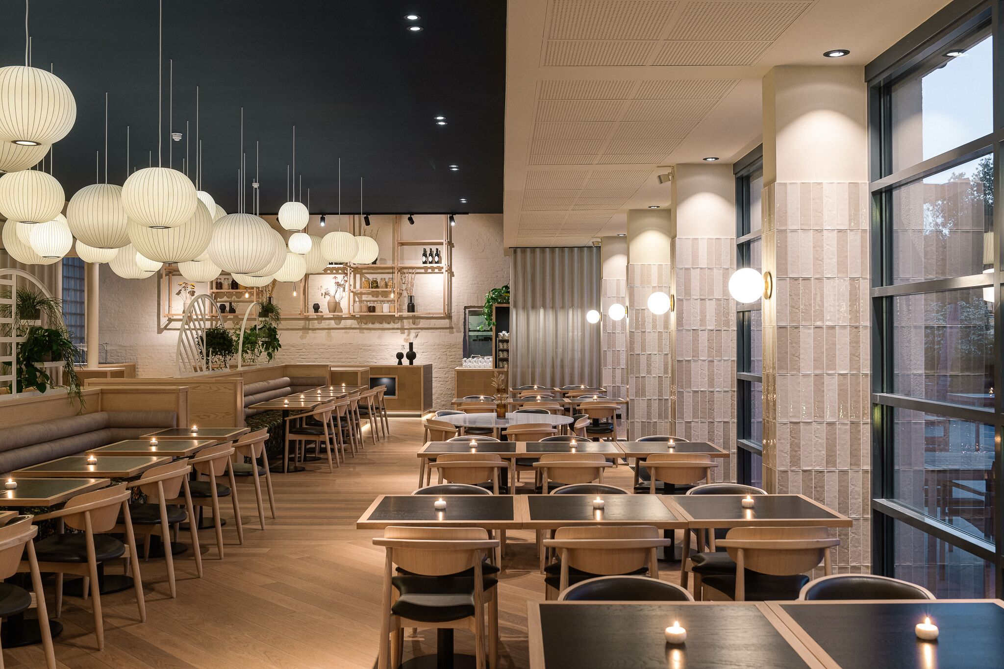 Een modern restaurantinterieur met houten tafels en stoelen, hangende pendellampen en uitzicht op het omliggende landschap door grote ramen.