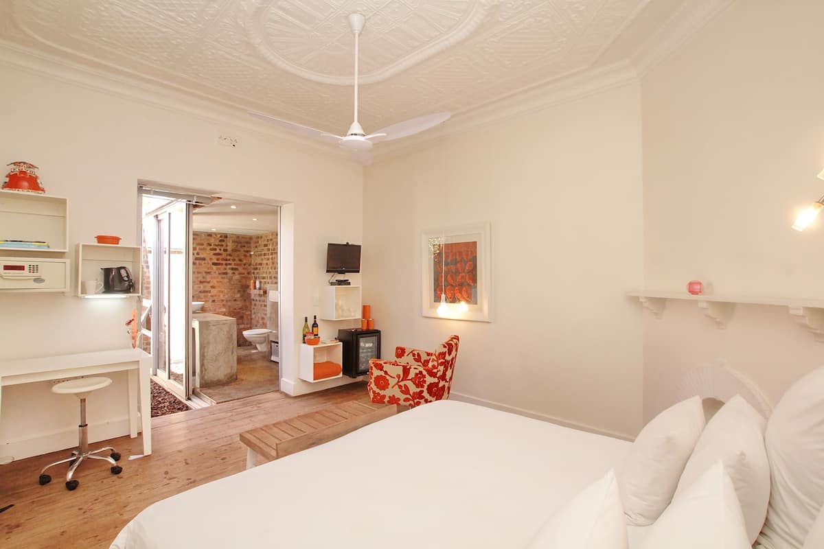 A bedroom with a bed, a desk, a chair, and a television. There is a door leading to a bathroom and a kitchen.