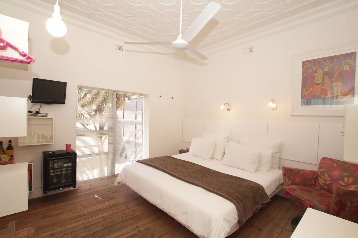 A bedroom with a bed, a ceiling fan, a television, a window with blinds, and a wall-mounted light fixture.