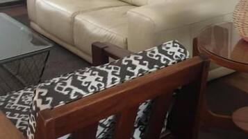 Una sala de estar con un sofá color beige, una silla de madera con cojines estampados, un aparador de madera y un cuadro en la pared.