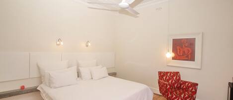 Une chambre avec un grand lit, un ventilateur de plafond, un fauteuil rouge et une œuvre d’art encadrée accrochée au mur.