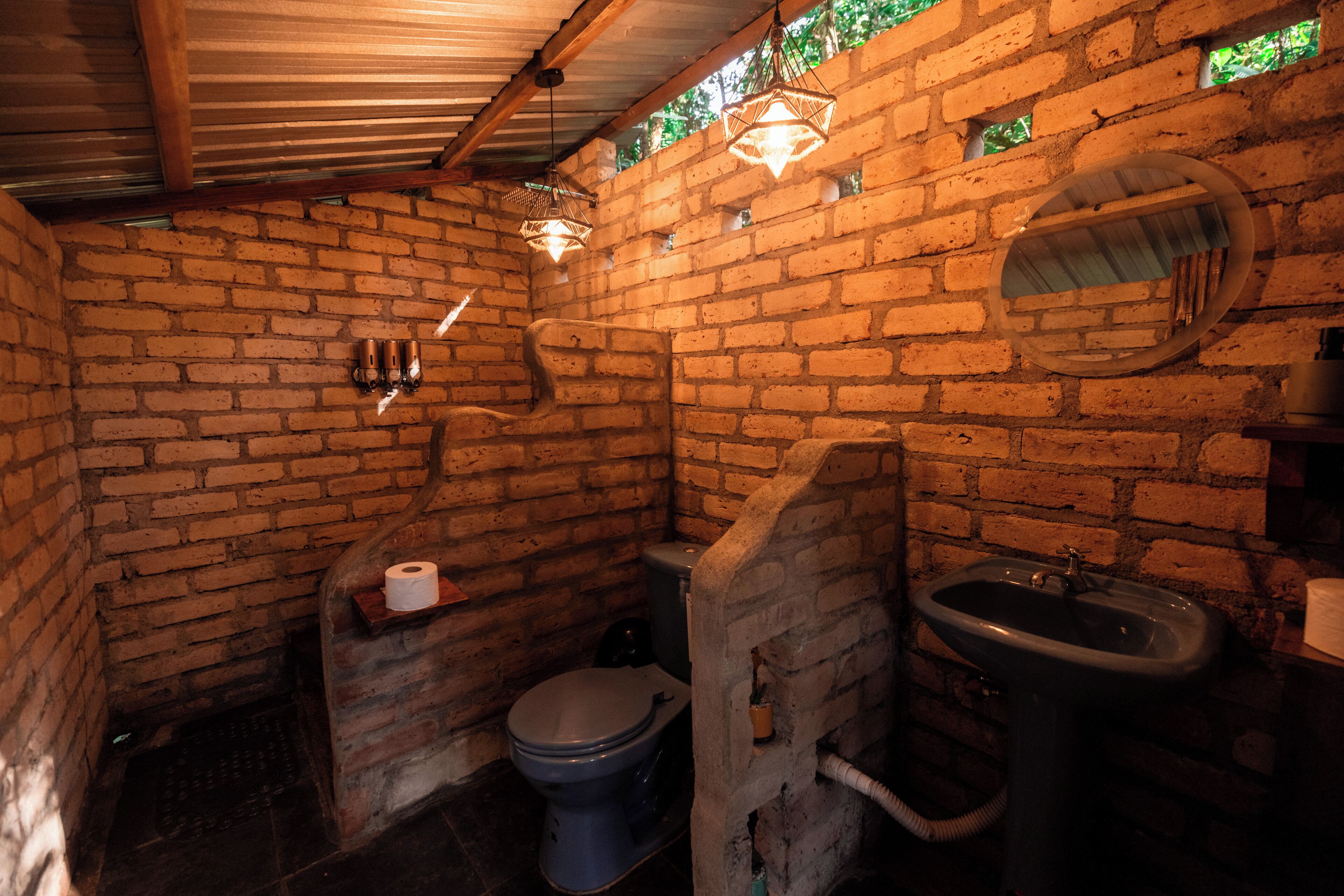 A rustic bathroom with exposed brick walls, a toilet, a sink, and hanging light fixtures.