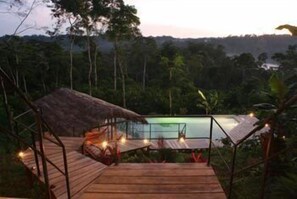 Una terraza de madera con techo de paja que da a una piscina, rodeada de exuberante vegetación tropical.