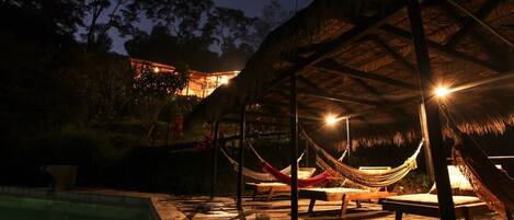 A nighttime outdoor area with a pool, hammocks, and a thatched-roof structure.