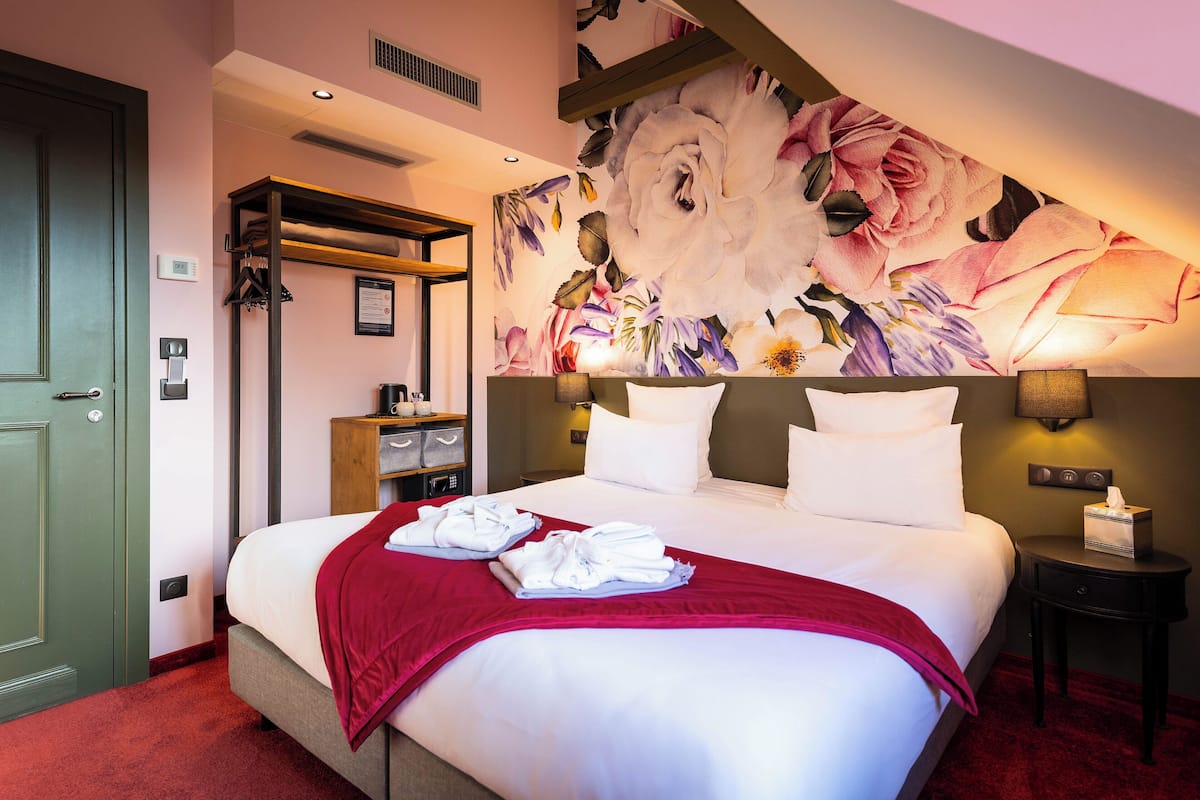 Une chambre d’hôtel avec un grand lit, un plaid rouge, des serviettes soigneusement pliées, une fresque murale à motifs floraux, une étagère en bois, une table de chevet avec une lampe et une petite table de nuit.