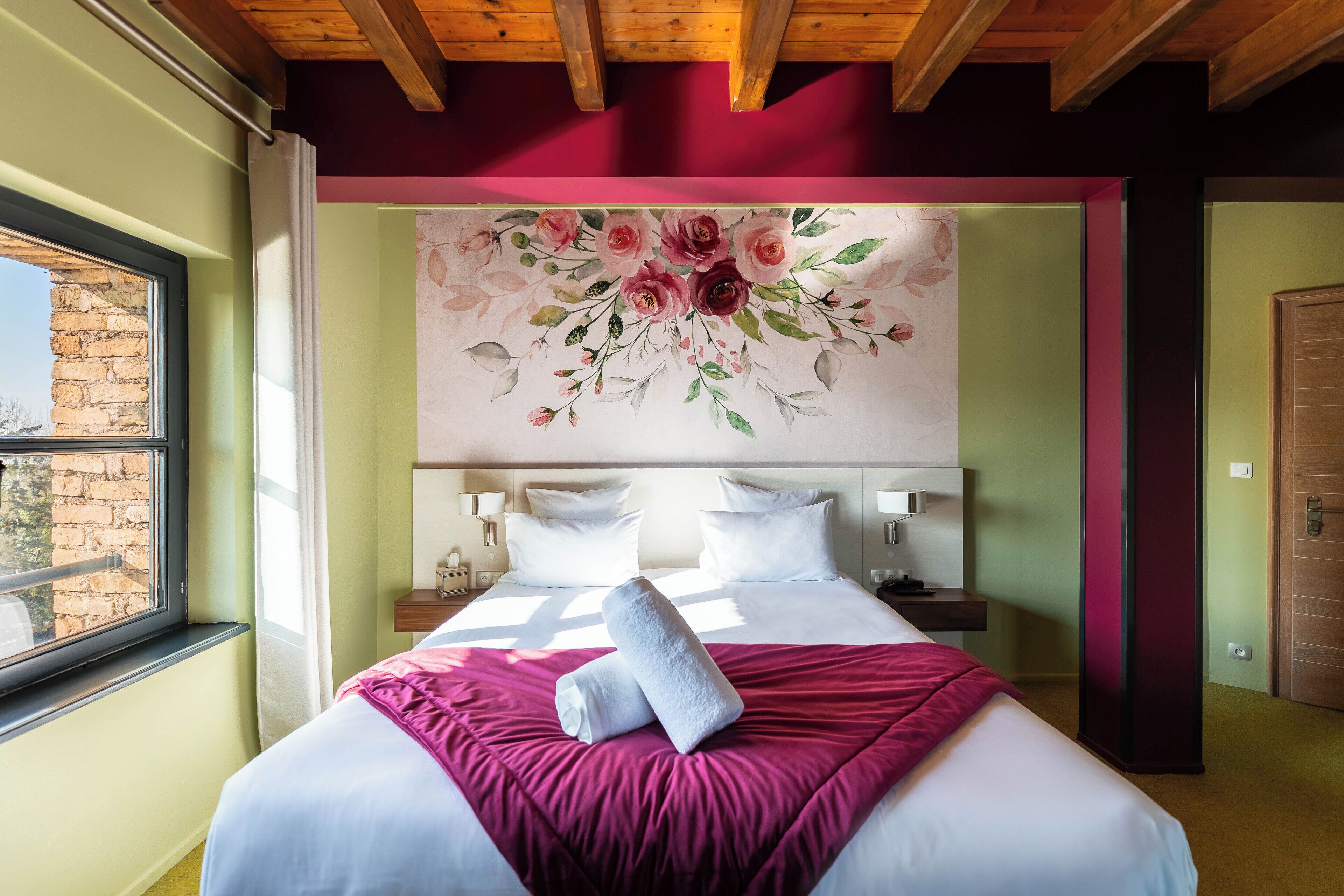 Habitación de hotel con una cama grande, un mural floral en la pared, una ventana que da a un muro de ladrillo y techo de madera.