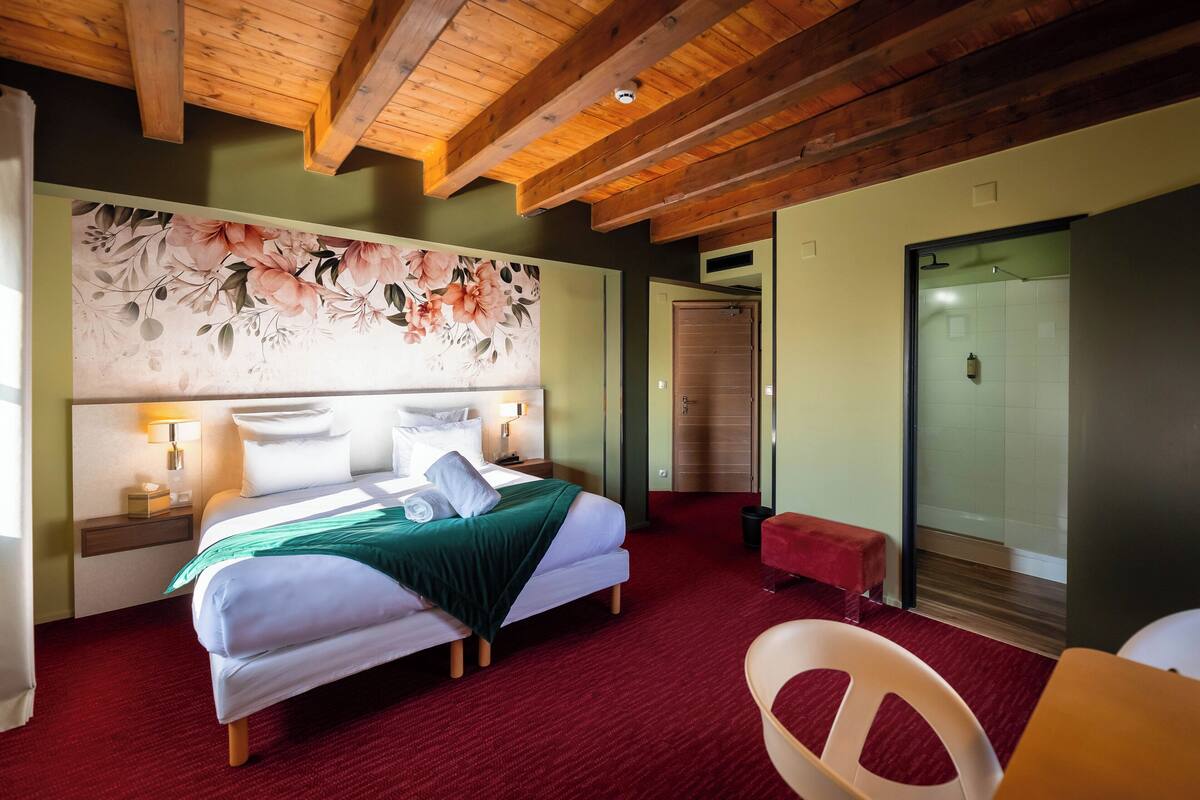Une chambre d’hôtel avec un plafond en bois, une grande tête de lit à motifs floraux, un lit avec du linge de maison blanc, une moquette rouge, un banc rouge et une chaise blanche.