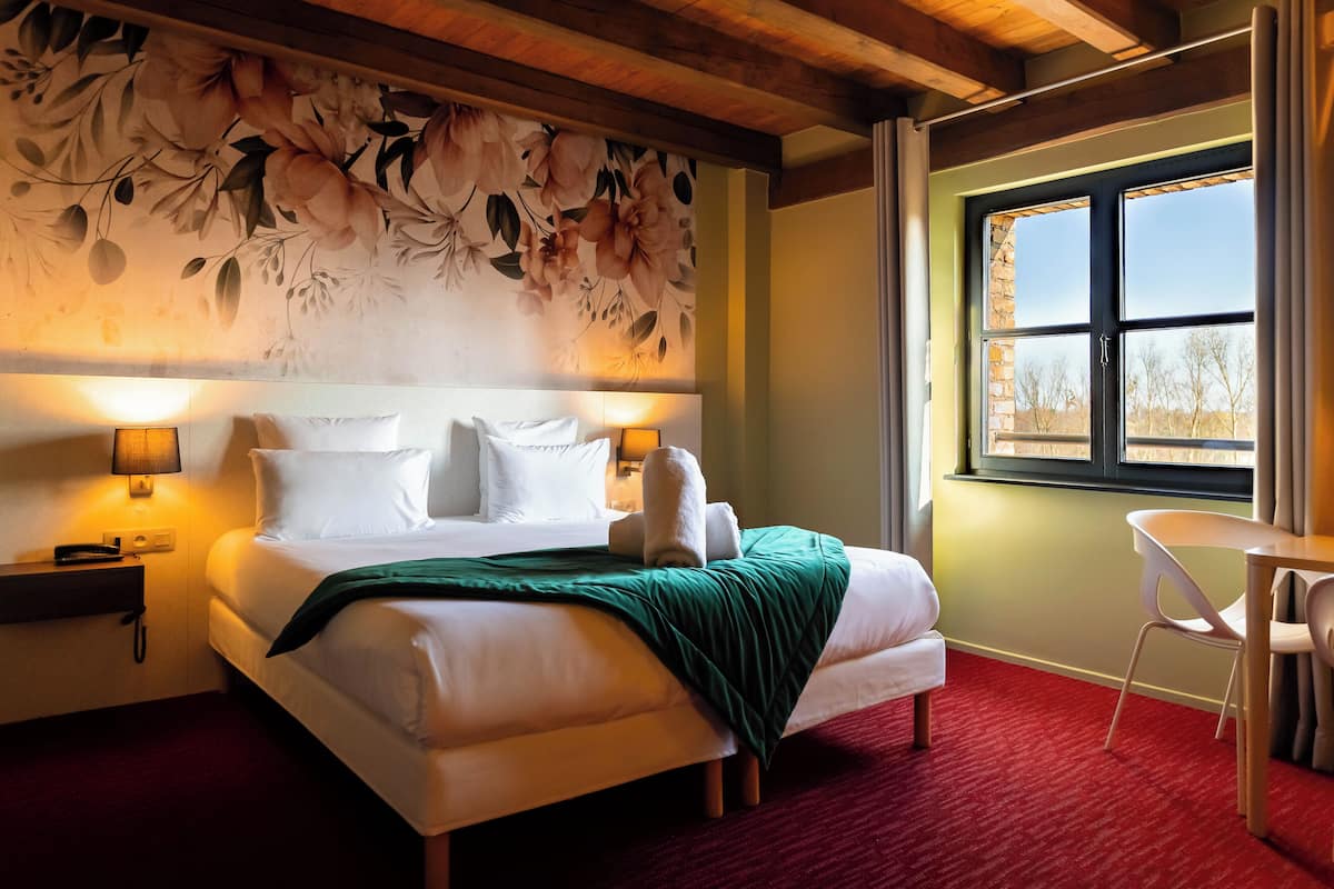 Une chambre d’hôtel avec un grand lit, un plaid vert, une tête de lit en bois, une table de chevet avec une lampe et une fenêtre donnant sur des arbres.