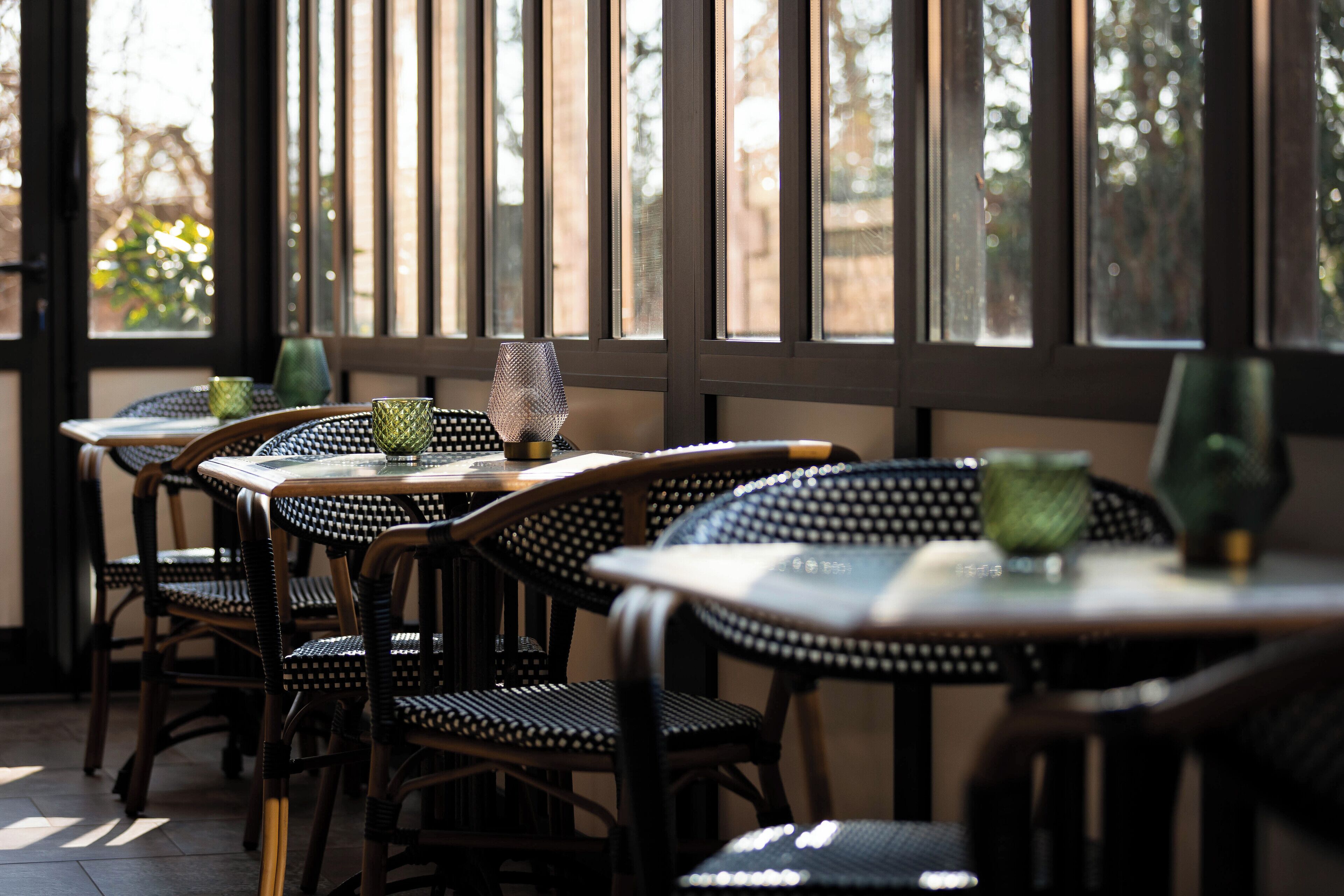 A restaurant interior with wicker chairs and tables, large windows, and a view of trees outside.