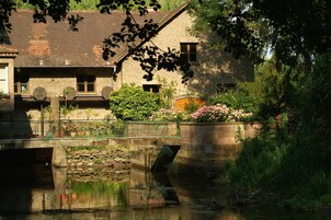 Un pont de pierre enjambe une rivière paisible, entouré d’une végétation luxuriante et bordé d’un bâtiment rustique à la façade de pierre et aux nombreuses fenêtres.