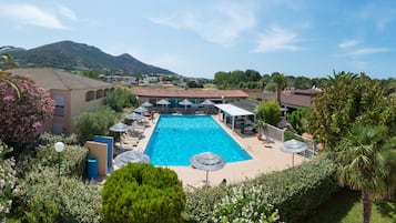 A swimming pool with lounge chairs and umbrellas, surrounded by greenery and buildings.
