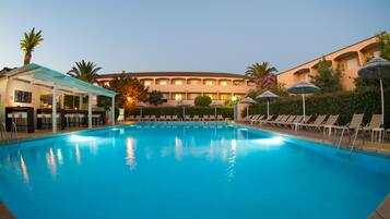 A swimming pool with lounge chairs and umbrellas, a covered seating area, and a building with multiple balconies.